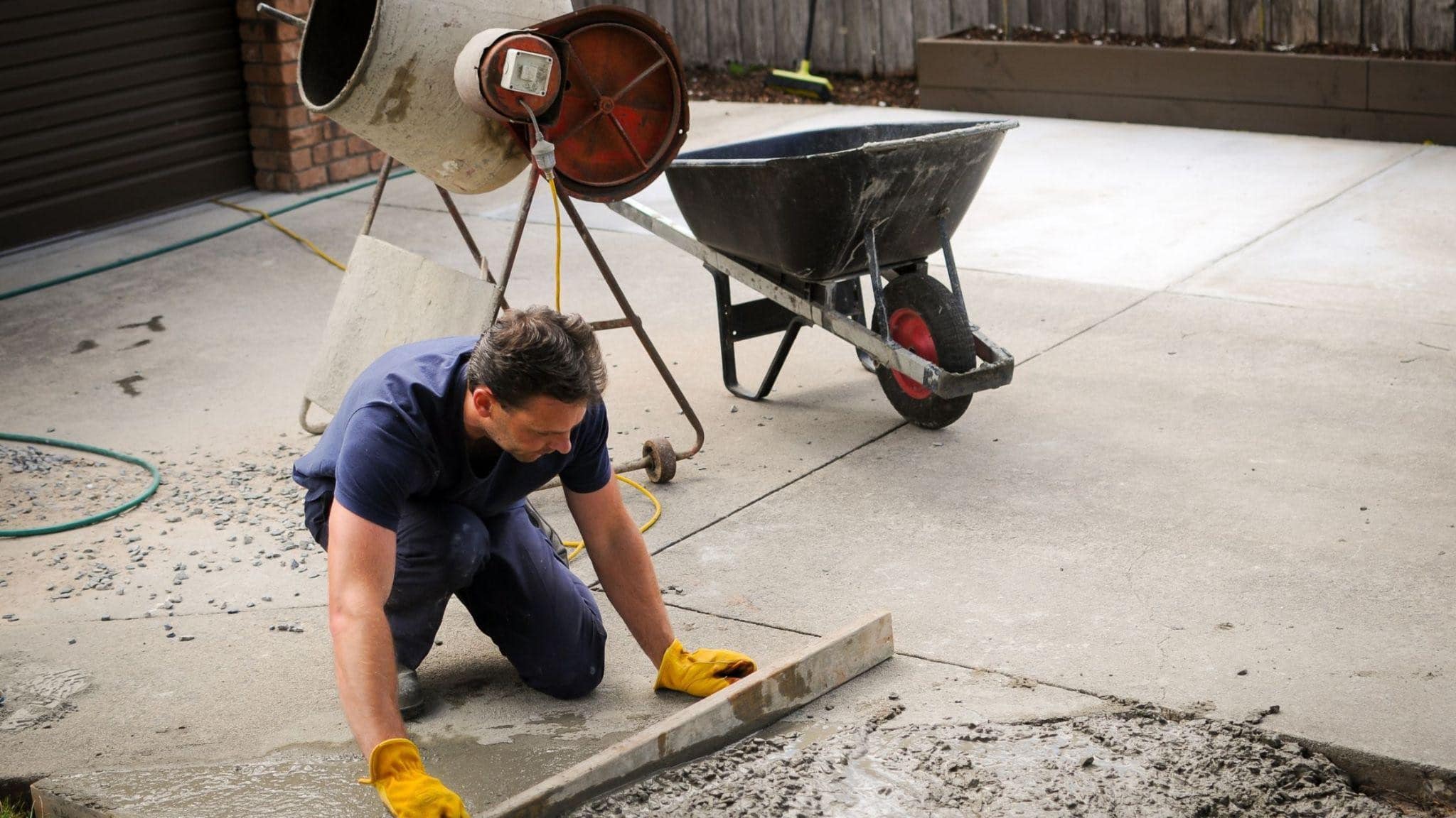 Worker smoothing wet concrete with a wooden screed on a driveway, with a cement mixer and wheelbarrow nearby
